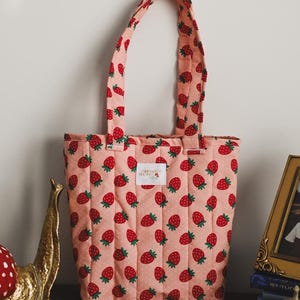 Könnte beinhalten: Eine rosafarbene gesteppte Tragetasche mit Erdbeer-Druck. Die Tasche hat ein weißes Etikett mit dem Text "Strawberry Garden Co."