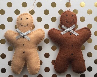 May include: Two felt gingerbread man ornaments, one brown and one beige, with silver ribbon bows. The ornaments are hanging on a white background with gold polka dots.