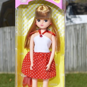 May include: A boxed doll with blonde hair wearing a red and white polka dot dress. The box is pink and yellow and has Japanese text on it.
