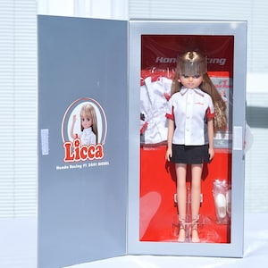 May include: A Licca doll in a display box. The doll is wearing a white shirt with red accents, a black skirt, and strappy shoes. The box is gray with a red interior and a Licca logo.