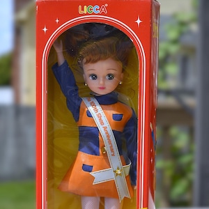 May include: A red box with a doll inside. The doll is wearing a blue and orange outfit with a white sash that says "100th Shop Anniversary". The box has the words "Licca" and "リカちゃん" on it.