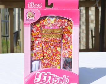 May include: A pink box containing a doll outfit. The outfit is a red kimono with a floral pattern, a gold obi, and black sandals. The box has the name "Licca" and Japanese text.