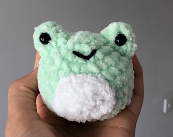 Pode incluir: Um pequeno brinquedo de pelúcia de sapo verde, feito de crochê com fio branco para a barriga. O sapo tem olhos pretos e um sorriso costurado.