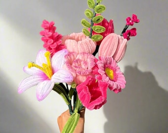 May include: A bouquet of pink and white pipe cleaner flowers. The flowers are arranged in a hand-held bouquet and include lilies, tulips, and daisies.