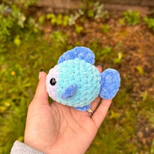 May include: A small crocheted blue and white fish with a single black eye. The fish is made with a fuzzy yarn and has a small fin on the back and a larger fin on the side.