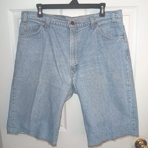 May include: Light blue denim shorts with a raw hem, hanging on a black hanger. The shorts have classic five-pocket styling, a button closure, and belt loops. The shorts are made of a light wash denim.