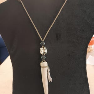 May include: A handcrafted necklace featuring a light-colored antler pendant. The pendant is adorned with black beads, patterned white beads, and a silver-colored charm. The necklace is suspended from a tan cord.