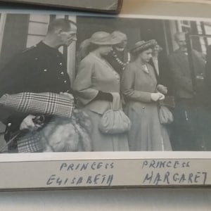 Puede incluir: Fotografía en blanco y negro de tres personas de pie frente a un edificio. Las personas visten atuendos formales. La foto está etiquetada como "Princesa Elizabeth" y "Princesa Margaret".