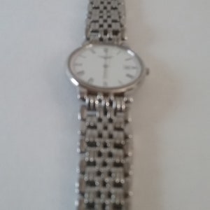 May include: Silver wristwatch with a white face and Roman numeral hour markers. The watch has a silver link band. The watch is displayed against a white background.
