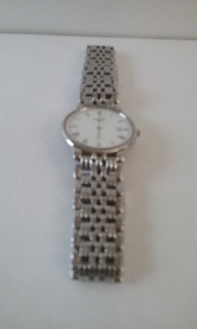May include: Silver wristwatch with a white face and Roman numeral hour markers. The watch has a silver link band. The watch is displayed against a white background.