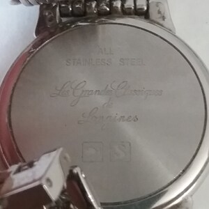 May include: Close-up of the back of a silver-toned watch. The watch face is engraved with the words "ALL STAINLESS STEEL" and "Les Grandes Classiques de Longines". The watch band is also silver-toned.