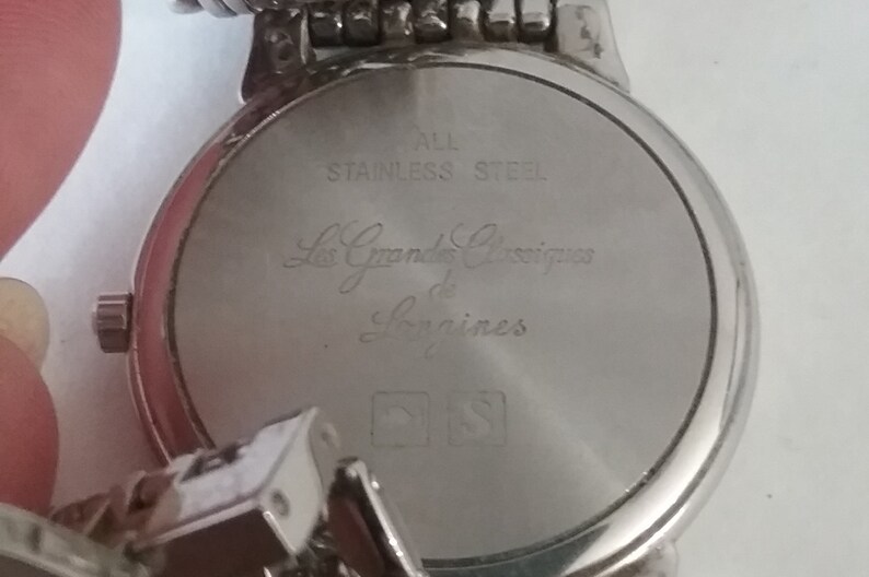 May include: Close-up of the back of a silver-toned watch. The watch face is engraved with the words "ALL STAINLESS STEEL" and "Les Grandes Classiques de Longines". The watch band is also silver-toned.