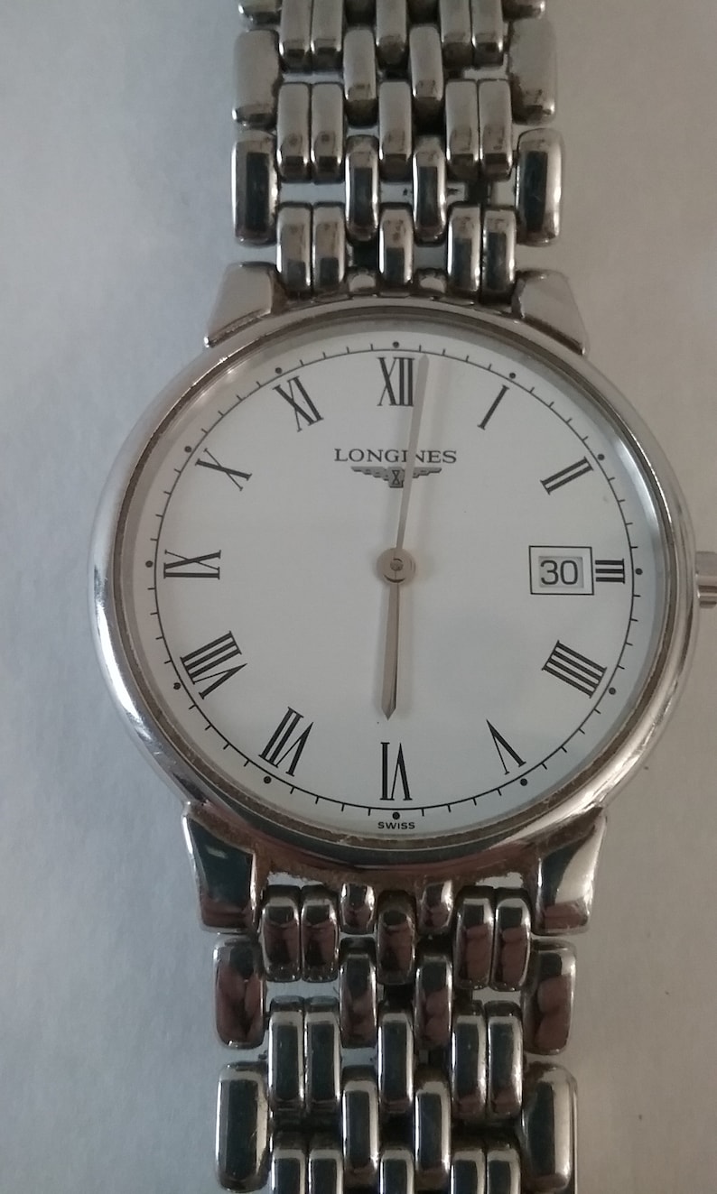 May include: A silver-toned Longines wristwatch with a white face, black Roman numerals, and a date display at the 3 o'clock position. The watch has a silver band and is marked "SWISS" at the bottom.