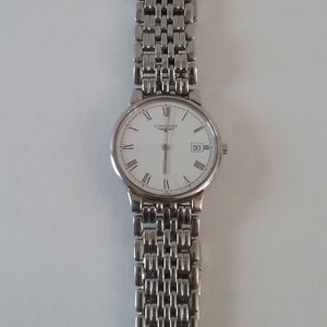 May include: A silver-toned wristwatch with a white face and Roman numeral hour markers. The watch has a date display at the 3 o'clock position and a linked metal band. The brand name "LONGINES" is printed on the watch face.