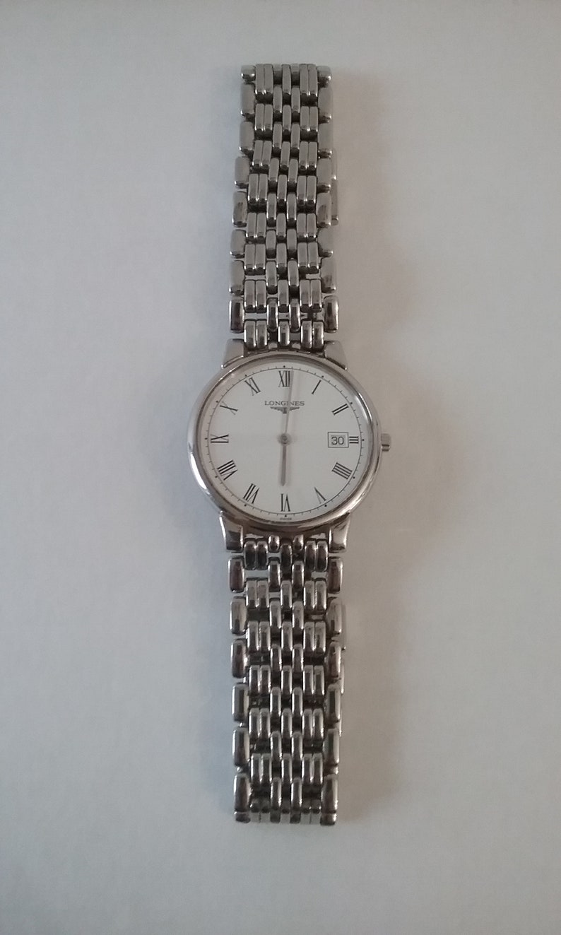 May include: A silver-toned wristwatch with a white face and Roman numeral hour markers. The watch has a date display at the 3 o'clock position and a linked metal band. The brand name "LONGINES" is printed on the watch face.
