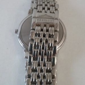 May include: A silver-toned wristwatch with a metal band. The watch face is round and silver, with a matching silver band. The band is made of small, rectangular links. The brand name "LONGINES" is visible on the clasp.