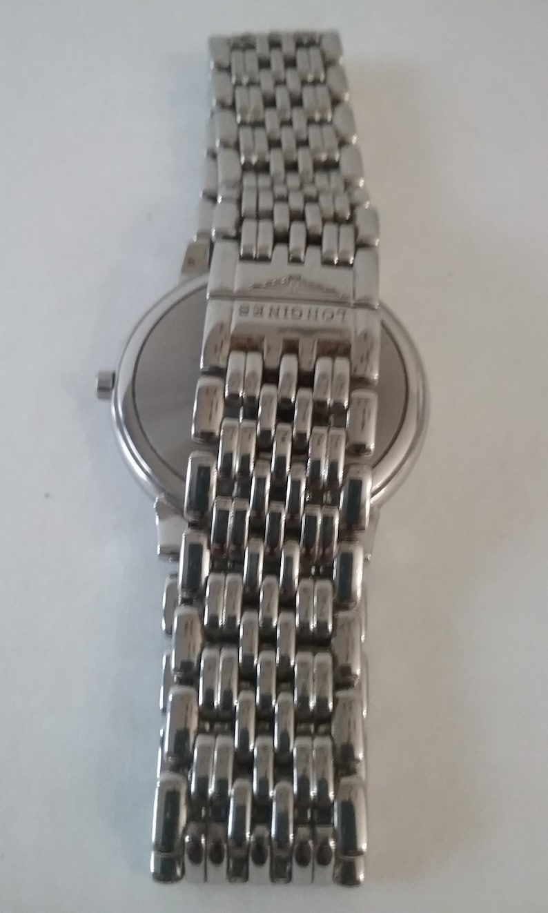 May include: A silver-toned wristwatch with a metal band. The watch face is round and silver, with a matching silver band. The band is made of small, rectangular links. The brand name "LONGINES" is visible on the clasp.