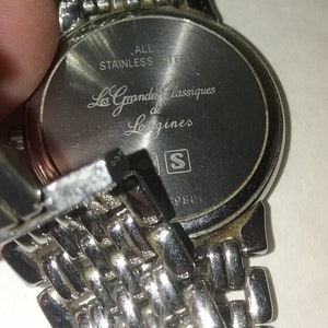 May include: Close-up of a stainless steel watch with a silver metal band. The watch face has the text "Les Grandes Classiques de Longines". The back of the watch is visible, with the words "ALL STAINLESS STEEL".