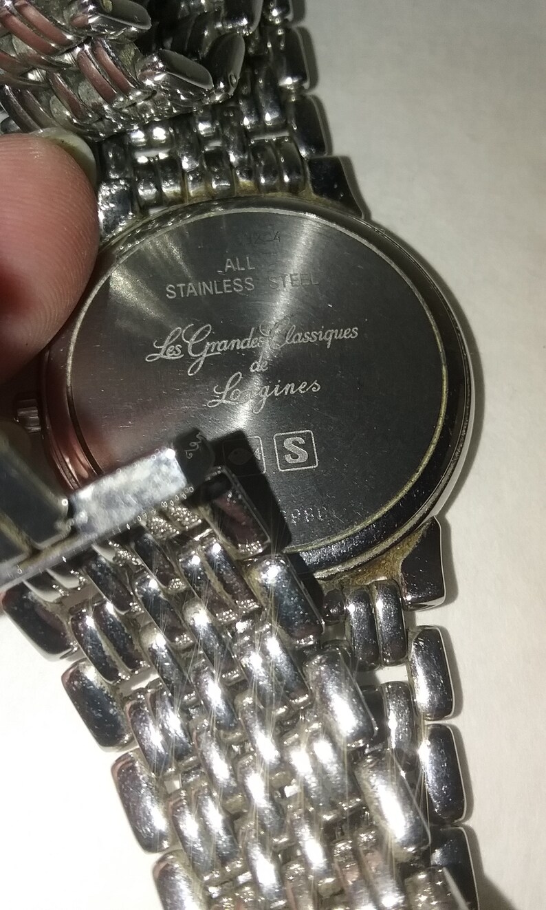 May include: Close-up of a stainless steel watch with a silver metal band. The watch face has the text "Les Grandes Classiques de Longines". The back of the watch is visible, with the words "ALL STAINLESS STEEL".