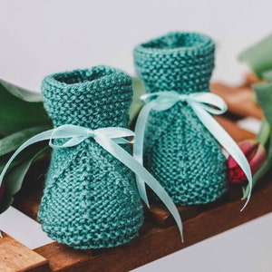 Könnte beinhalten: Zwei türkisfarbene gestrickte Babyschuhe mit hellblauen Satinbändern. Die Schuhe liegen auf einer Holzoberfläche mit roten Tulpen im Hintergrund.