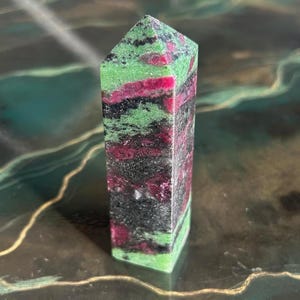 May include: A rectangular, polished gemstone obelisk. The stone features layers of green, deep red, black, and grey. The obelisk has a flat base and a pointed top. The gemstone is displayed on a reflective, dark green surface.