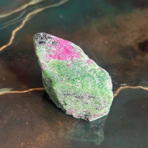 Könnte beinhalten: Ein roh geschliffener Edelstein mit einer lebendigen Farbpalette. Der Stein weist ein auffälliges Band in leuchtendem Pink auf, das mit Bereichen in sattem Grün kontrastiert und mit Schwarz gesprenkelt ist. Die Form ist unregelmäßig und natürlich.