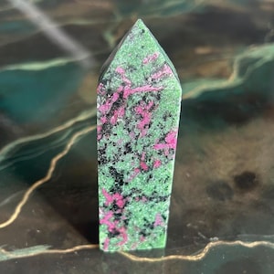 May include: A rectangular gemstone with a pointed top, polished to a smooth finish. The stone's base is green, with black and pink inclusions. This natural mineral is likely used for decorative or spiritual purposes, with a height of approximately 7 cm.