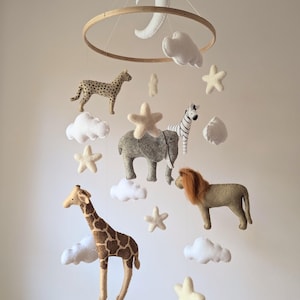Puede incluir: Un móvil de bebé de madera con una luna de fieltro blanco, nubes y estrellas. El móvil también presenta figuras de animales de fieltro, incluyendo una jirafa, un león, un elefante, una cebra y un guepardo.