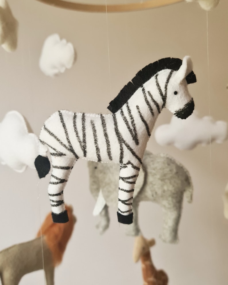May include: A black and white felt zebra hanging from a mobile with other felt animals and clouds. The zebra has a black mane and black hooves.