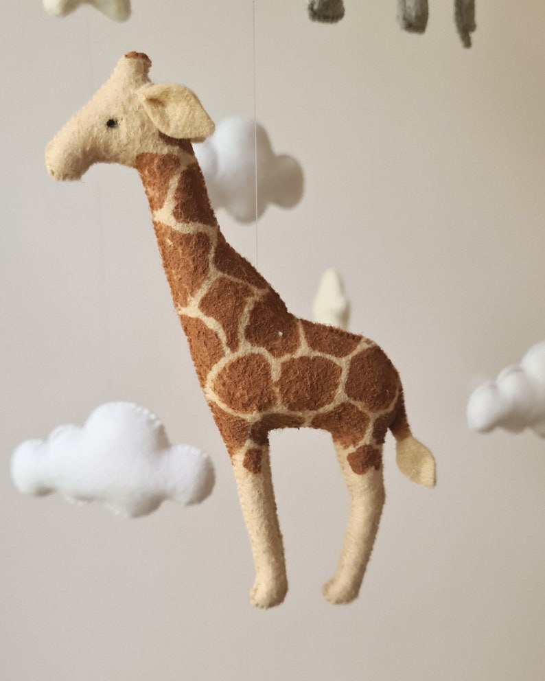 May include: A felt giraffe mobile hanging from a white ceiling. The giraffe is brown with a tan face and legs. The giraffe is surrounded by white felt clouds.
