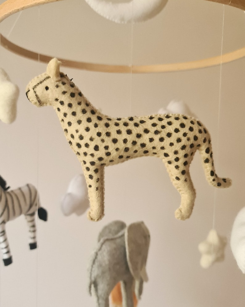 May include: A beige and black cheetah plush toy hangs from a wooden mobile. The cheetah has black spots and is made of felt.
