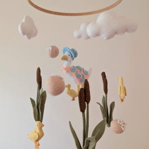 Pode incluir: Móvel de bebê artesanal com patos de feltro, nuvens, juncos e um ganso usando um chapéu e xale. O móvel é suspenso por um aro de madeira. A paleta de cores inclui branco, amarelo, rosa, azul, marrom e verde.