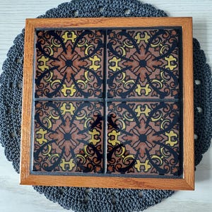 May include: A square wooden framed tile trivet with four brown, black, and yellow patterned tiles.