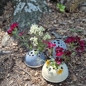 May include: Three dome-shaped ceramic flower frogs with multiple circular openings for stems. Two are glazed in a glossy blue finish, and one is in a matte cream finish. Each holds a variety of fresh red, white, and yellow wildflowers.