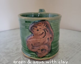 May include: Green and aqua ceramic mug with a clay dragon design.