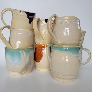 May include: A collection of handmade ceramic mugs in various sizes and designs. The mugs feature a creamy white base with accents of blue, turquoise, and orange glaze. Each mug has a unique handle and shape, showcasing the artistry of the pottery.