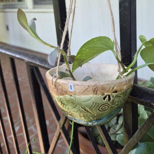 May include: A ceramic hanging planter with a green and tan glazed finish featuring embossed spiral and leaf patterns. The bowl is suspended by natural twine and contains a green leafy plant. A small white sticker with the number 1 is attached.
