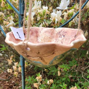 May include: A handmade, ceramic bird feeder with a scalloped edge, suspended by twine. The feeder has a pinkish-brown interior with floral patterns and a turquoise rim. A small white tag with the number "1" is attached.