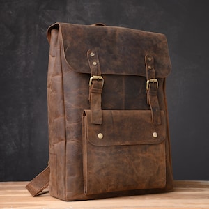 Op de afbeelding: Een bruine leren rugzak met een groot voorvak en twee verstelbare banden. De rugzak heeft een verweerde look en is gemaakt van hoogwaardig leer.