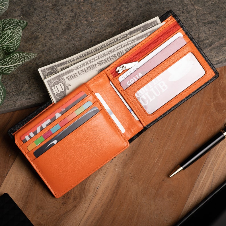 May include: A black leather wallet with an orange interior. The wallet is open and has several card slots, a billfold, and a clear ID window. The wallet is lying on a wooden surface with some US dollar bills and a pen.
