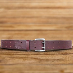 May include: A brown leather belt with a silver buckle and a perforated design. The belt is on a wooden surface.