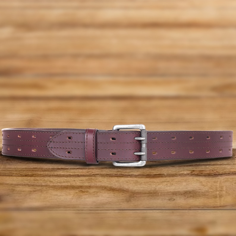 May include: A brown leather belt with a silver buckle and a perforated design. The belt is on a wooden surface.