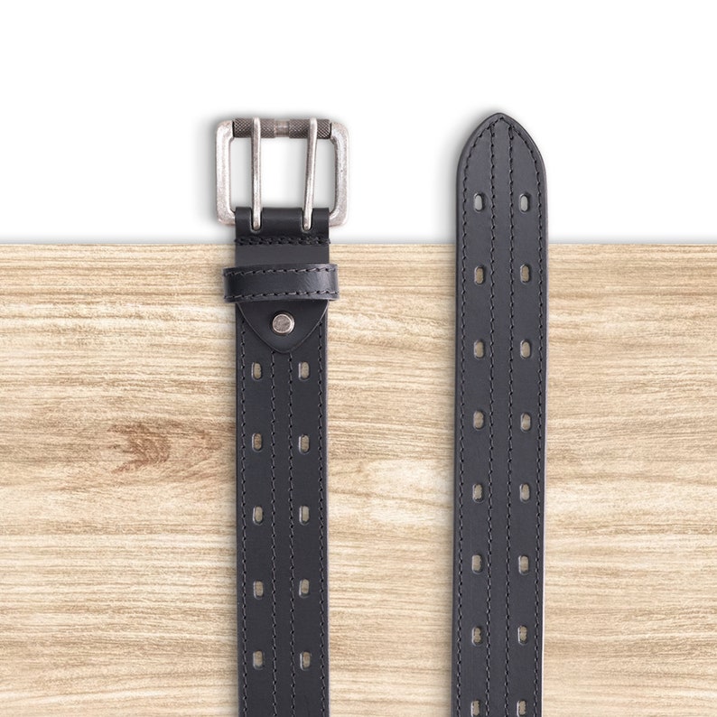 May include: A black leather belt with a silver buckle and multiple rows of stitching. The belt is on a wooden surface.