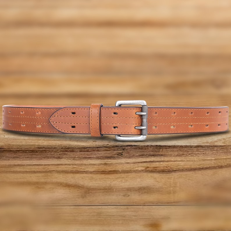 May include: A brown leather belt with a silver buckle and a perforated design. The belt is on a wooden surface.