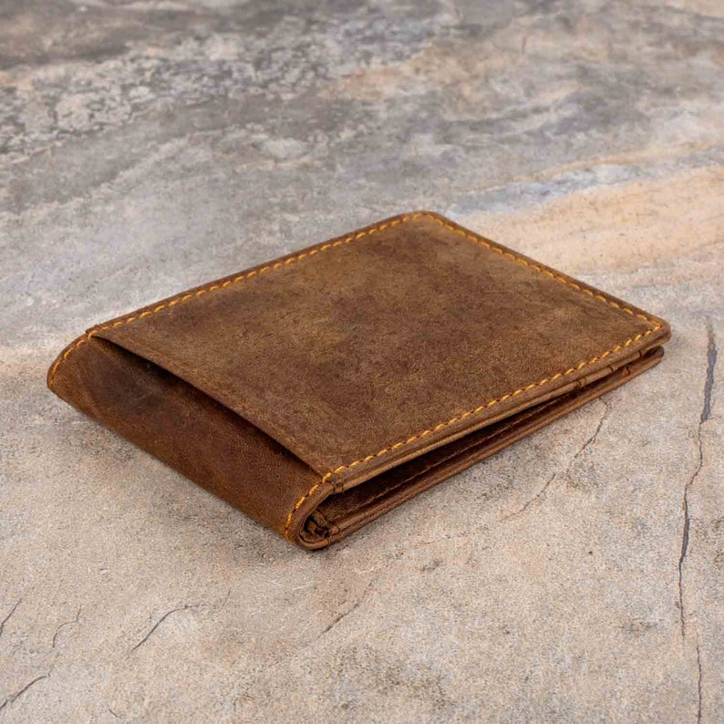 May include: A brown leather wallet with yellow stitching. The wallet is folded closed and lying on a gray textured surface.