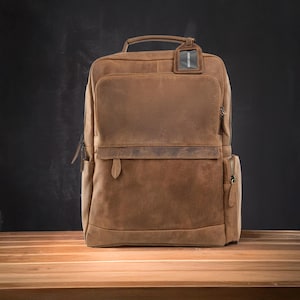 Op de afbeelding: Een bruine leren rugzak met meerdere vakken en een handvat aan de bovenkant. De tas heeft een rechthoekige vorm met een klep aan de voorkant en een klein label aan het handvat. De rugzak staat op een houten oppervlak.