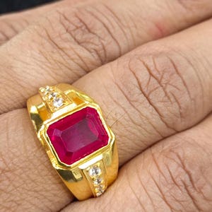 Peut inclure: Une bague en or ornée d'une grande pierre précieuse rectangulaire rouge foncé au centre. L'anneau est fabriqué en or jaune poli et comprend deux rangées de petites pierres d'accent claires et scintillantes incrustées sur les côtés.