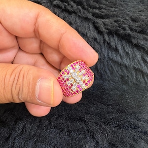 May include: A gold ring with a floral design. The ring is encrusted with small, round, pink and white gemstones. The gemstones are arranged in a pattern that resembles a flower. The ring is held in a hand against a black background.