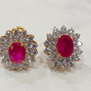 Könnte beinhalten: Ohrstecker-Paar mit einem ovalen, roten Edelstein, umgeben von einem Kranz aus klaren Steinen. Die Ohrringe sind in einem goldfarbenen Metall gefasst, was einen schönen Kontrast erzeugt.