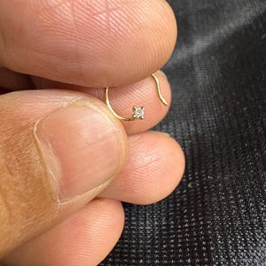 Peut inclure: Une petite boucle d'oreille en or délicate avec une seule pierre précieuse claire taillée en carré. La boucle d'oreille est tenue entre les doigts sur un fond sombre, mettant en valeur ses détails fins et son design minimaliste.